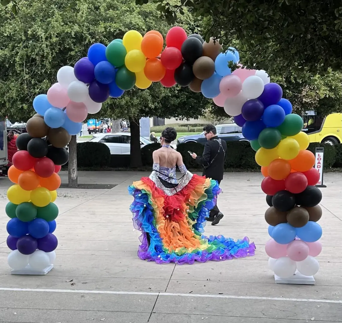 Person wearing rainbow ruffled gown under balloon arch